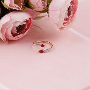 May include: A gold-toned open ring with a row of small, clear stones and two teardrop-shaped red gemstones. The ring is displayed on a pink surface, with pink flowers in the background.