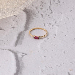 Może przedstawiać: Złoty pierścionek z prostokątnym, głęboko czerwonym kamieniem, otoczonym małymi, przezroczystymi kamieniami. Pierścionek jest umieszczony na marmurowej, jasnej powierzchni, z delikatnym liściem w tle. Prosta i elegancka biżuteria.