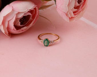 Puede incluir: Un anillo de oro con una piedra central de esmeralda ovalada y pequeñas piedras de acento. El anillo está sobre un fondo rosa con flores rosas en el fondo. El anillo es una joya.