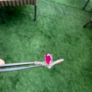 Pu&ograve; includere: Un anello d'oro con una gemma rossa brillante a forma di pera. L'anello ha un design a forma di V con piccole gemme trasparenti. &Egrave; tenuto da una pinzetta su uno sfondo verde.