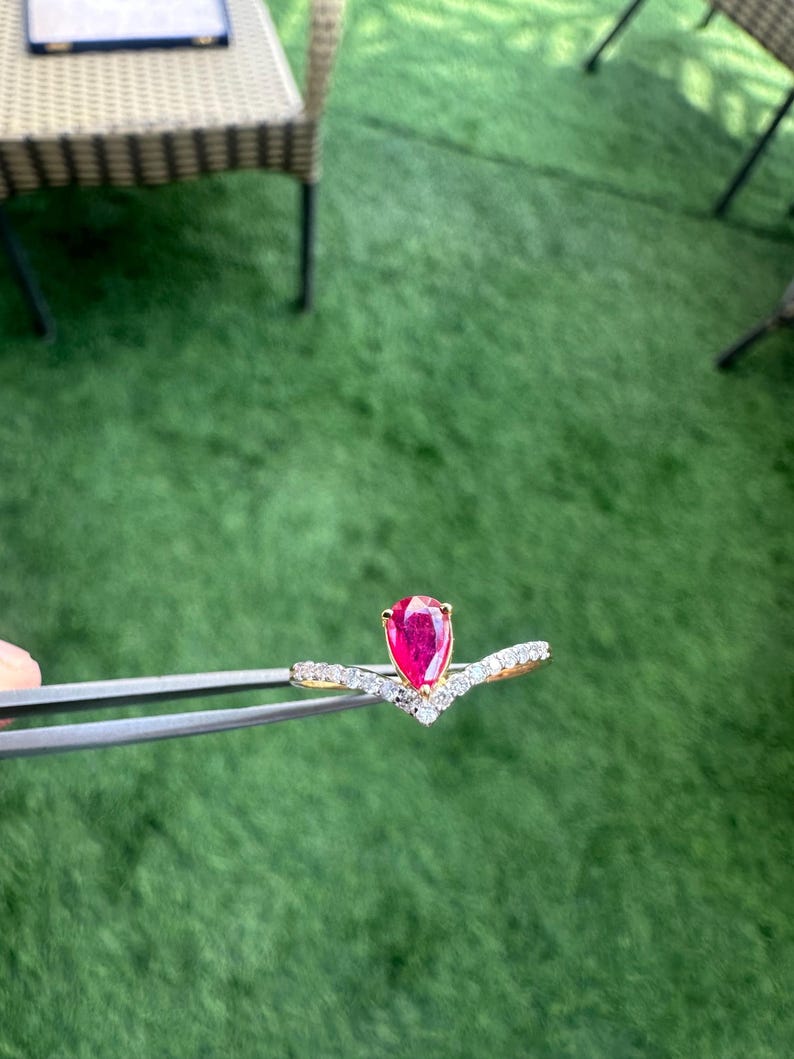 Pu&ograve; includere: Un anello d'oro con una gemma rossa brillante a forma di pera. L'anello ha un design a forma di V con piccole gemme trasparenti. &Egrave; tenuto da una pinzetta su uno sfondo verde.