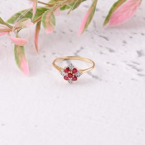 Pode incluir: Um anel de ouro com um design floral, com pedras preciosas vermelhas no centro e pedras preciosas brancas nos cantos. O anel está sobre um fundo branco com folhagem rosa e verde.