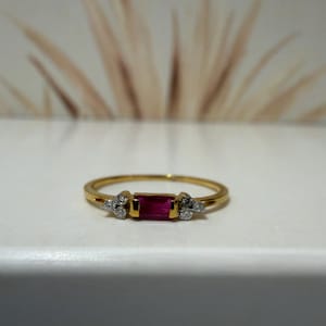 May include: A gold ring featuring a central rectangular baguette-cut red gemstone. The red stone is flanked on both sides by a cluster of three small, round, clear white stones set into the gold band, creating a delicate and balanced design.