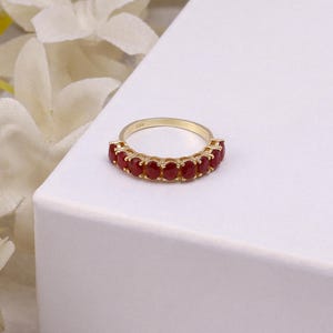 Op de afbeelding: Een gouden ring met een rij rode edelstenen. De ring heeft een gepolijste gouden band en de rode edelstenen zijn dicht op elkaar geplaatst. De ring wordt gepresenteerd op een wit oppervlak, waardoor het ontwerp en de kleur worden benadrukt.