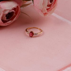 Puede incluir: Anillo dorado con una gran gema de rubí ovalada en el centro, flanqueada por piedras blancas más pequeñas. El anillo está sobre un fondo rosa con flores rosas en la esquina superior izquierda.