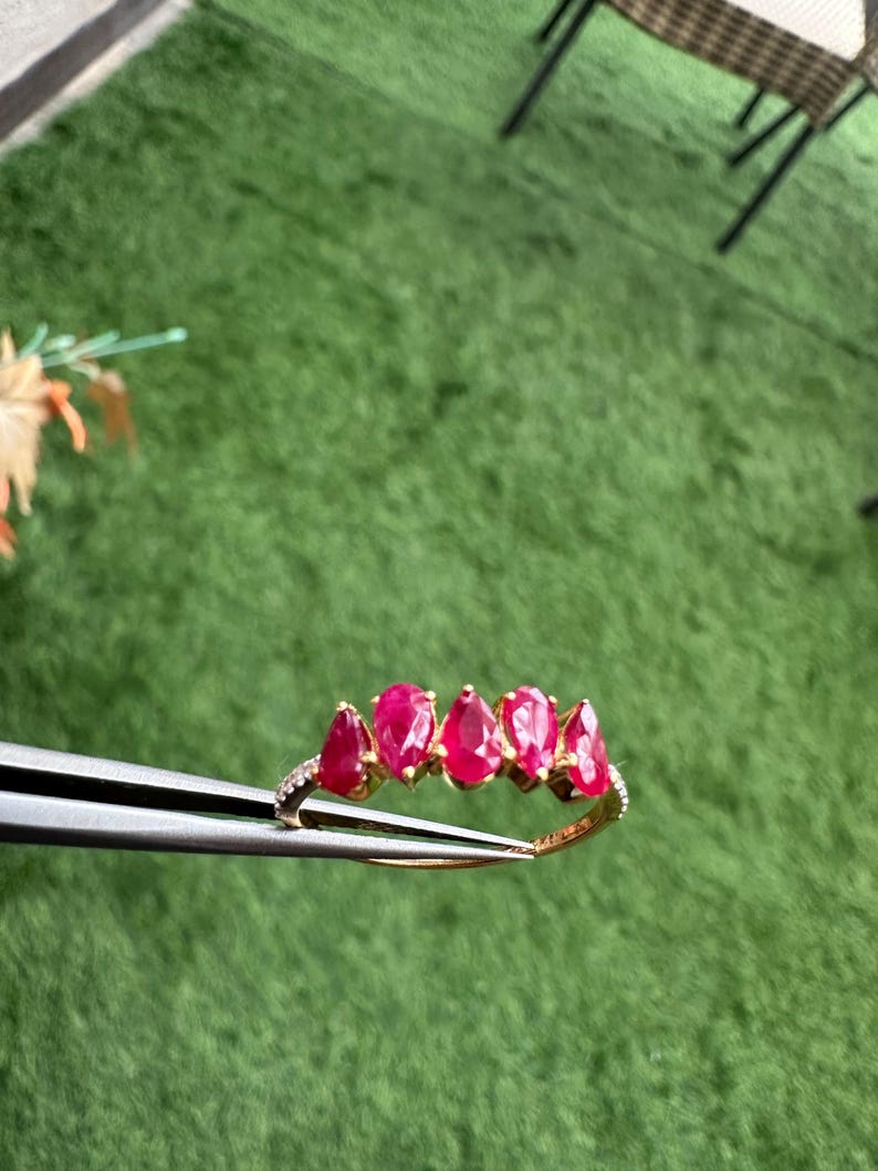 Pode incluir: Um anel de ouro com cinco pedras preciosas vermelhas vibrantes em forma de p&ecirc;ra. O anel &eacute; adornado com pequenas pedras transparentes ao longo da banda. O anel &eacute; segurado por uma pin&ccedil;a contra um fundo verde.