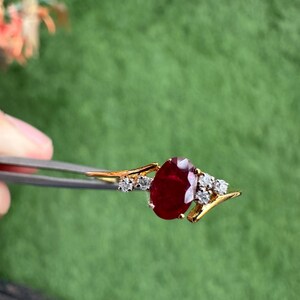 May include: A gold ring with a large, oval-cut ruby gemstone. The ruby is a deep red colour and is accented by several small, clear diamonds. The ring is held by tweezers against a blurred green background.