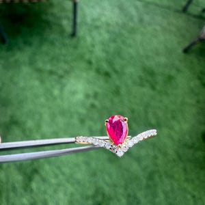 Pu&ograve; includere: Un anello d'oro con una gemma rossa a forma di pera al centro. La fascia dell'anello &egrave; ornata da piccole gemme bianche scintillanti, creando un design a forma di V. L'anello &egrave; tenuto da una pinzetta su uno sfondo verde.