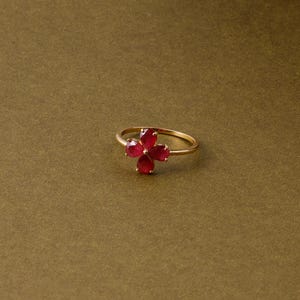 May include: A gold ring with a four-petal flower design. Each petal is a vibrant red gemstone. The ring is set against a solid, olive-green background, highlighting the jewelry's details.