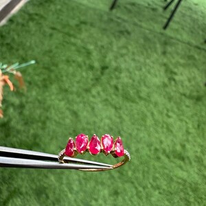 Pode incluir: Um anel de ouro com cinco rubis em forma de gota. O anel &eacute; adornado com pequenas pedras preciosas transparentes ao longo da banda. O anel &eacute; segurado por uma pin&ccedil;a contra um fundo verde.