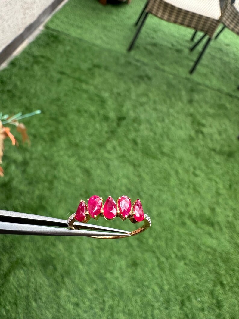 Pode incluir: Um anel de ouro com cinco rubis em forma de gota. O anel &eacute; adornado com pequenas pedras preciosas transparentes ao longo da banda. O anel &eacute; segurado por uma pin&ccedil;a contra um fundo verde.