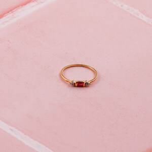 May include: A gold ring featuring a rectangular red gemstone, with two smaller clear stones either side. The ring is set on a pink tiled surface. The design is simple and elegant.