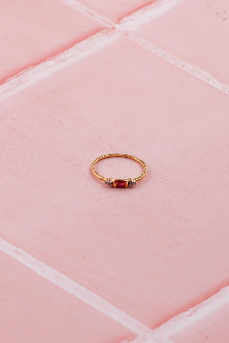 May include: A gold ring featuring a rectangular red gemstone, with two smaller clear stones either side. The ring is set on a pink tiled surface. The design is simple and elegant.