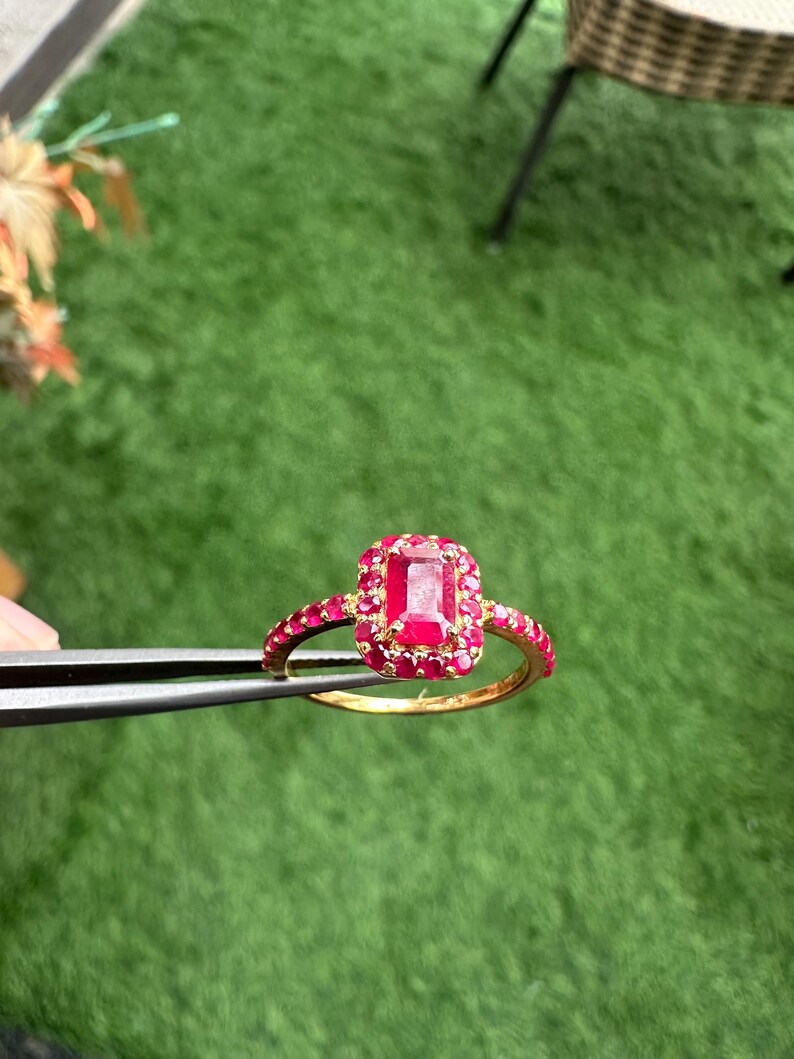 以下が含まれることがあります： 中央に大きな長方形の赤い宝石、周囲に小さな赤い宝石がちりばめられたゴールドリング。リングのバンドにも赤い宝石が飾られています。リングは、緑色の背景に対してピンセットで保持されています。