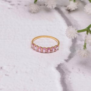Puede incluir: Un anillo de oro con una serie de piedras preciosas rosas de corte ovalado. El anillo está colocado sobre una superficie blanca y gris con aspecto de mármol, con delicadas flores blancas en el fondo. Es una joya.