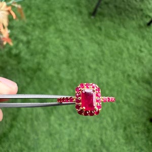 以下が含まれることがあります： 大きな長方形のルビーのセンター石と、小さな丸いルビーに囲まれた金の指輪。指輪のバンドにもルビーが飾られています。指輪は緑色の背景に対してピンセットで保持されています。