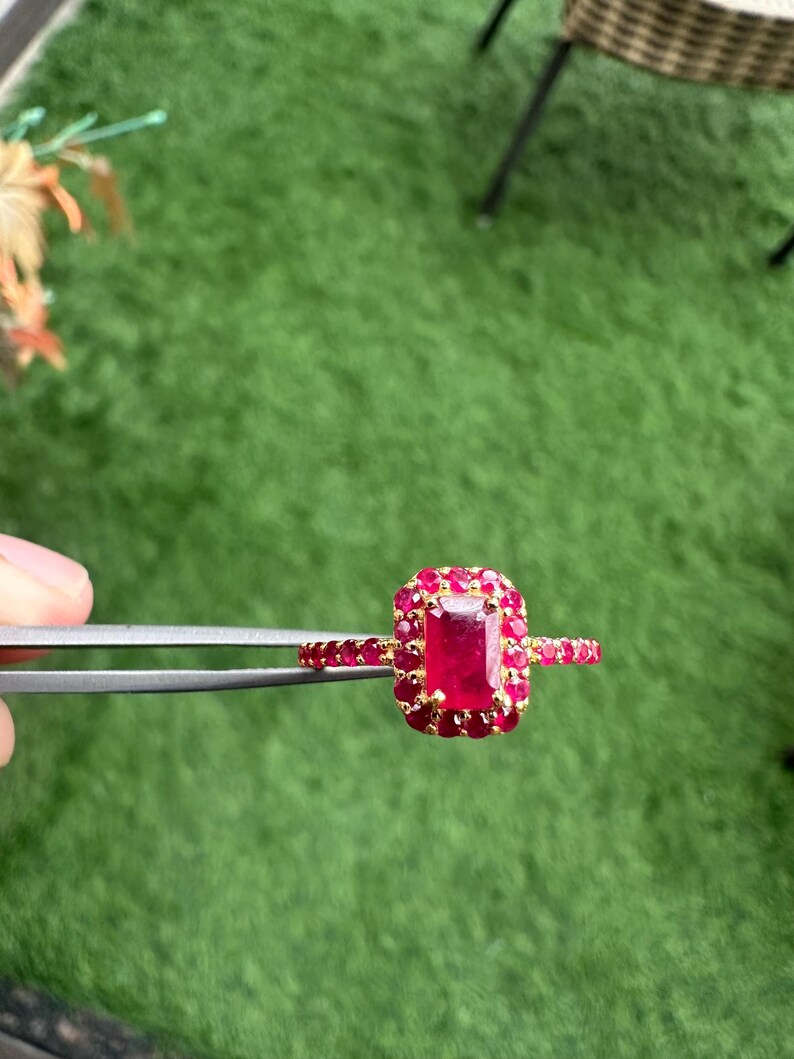 以下が含まれることがあります： 大きな長方形のルビーのセンター石と、小さな丸いルビーに囲まれた金の指輪。指輪のバンドにもルビーが飾られています。指輪は緑色の背景に対してピンセットで保持されています。