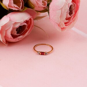 May include: A gold ring featuring a rectangular red gemstone and two small white gemstones. The ring is set against a pink backdrop, with pink flowers in the background. A delicate piece of jewellery.