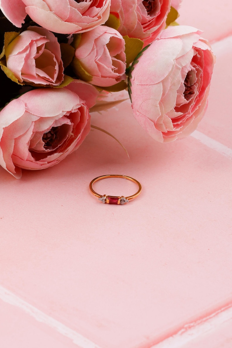 May include: A gold ring featuring a rectangular red gemstone and two small white gemstones. The ring is set against a pink backdrop, with pink flowers in the background. A delicate piece of jewellery.