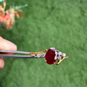 May include: A gold ring with a large, oval ruby gemstone and smaller diamonds. The ring is held by tweezers against a green background. The ruby is the focal point of the ring.