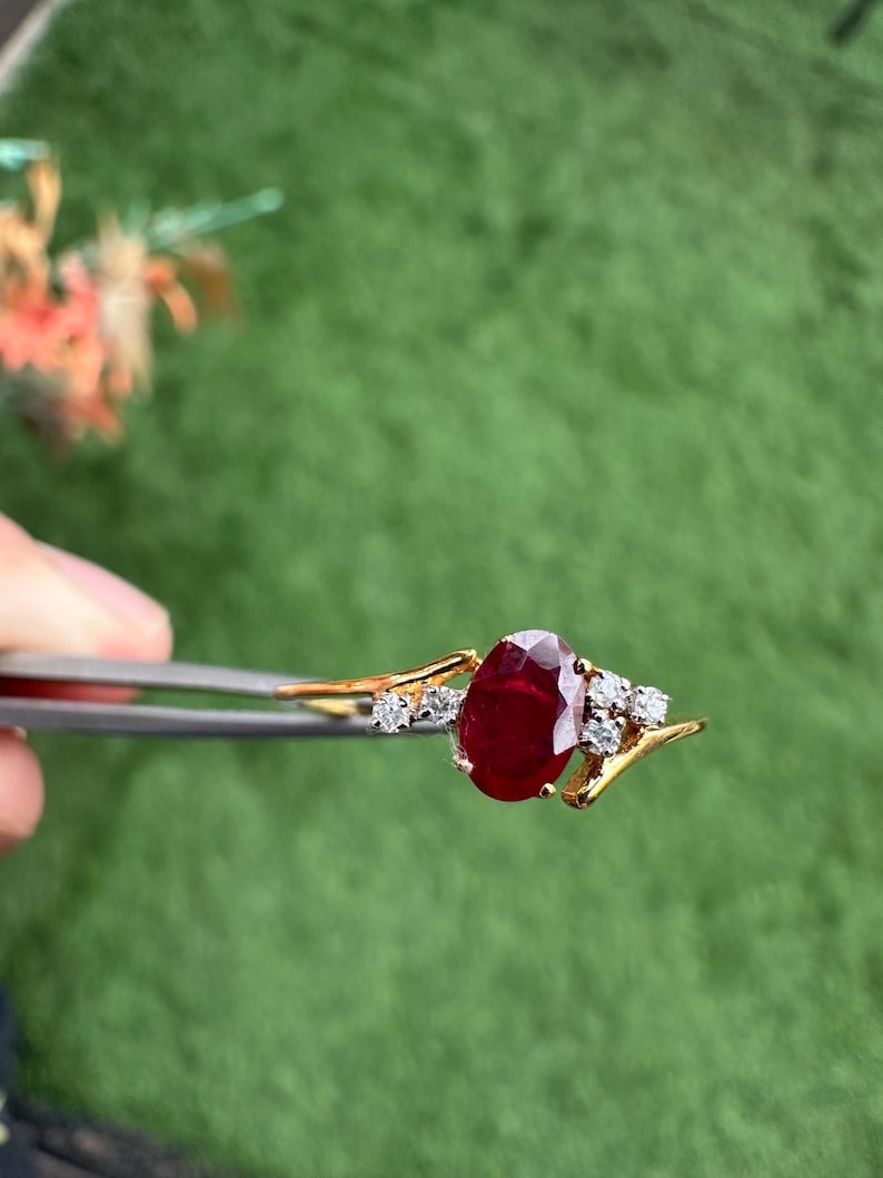 May include: A gold ring with a large, oval ruby gemstone and smaller diamonds. The ring is held by tweezers against a green background. The ruby is the focal point of the ring.