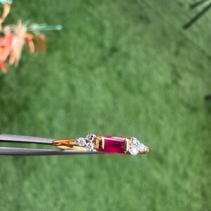 May include: A gold ring with a rectangular, deep red gemstone and clusters of small, clear gemstones. The ring is held by tweezers against a green background.