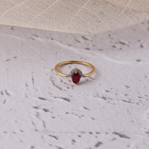 Op de afbeelding: Een gouden ring met een ovaalvormige, dieprode edelsteen in het midden. De ring heeft een delicaat ontwerp met kleine, heldere accentstenen rond de hoofdsteen. De ring is geplaatst tegen een neutrale achtergrond.