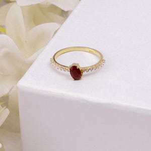 Op de afbeelding: Een gouden ring met een centraal ovaalvormig, dieprood edelsteen. De band is versierd met kleine, heldere edelstenen. De ring staat tegen een witte achtergrond, met zachte, witte bloemen op de achtergrond.