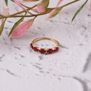 Puede incluir: Un anillo de oro con piedras preciosas rojas oscuras de corte ovalado. El anillo se encuentra sobre una superficie blanca y gris con efecto mármol, con follaje rosa y verde en el fondo. Es una joya.