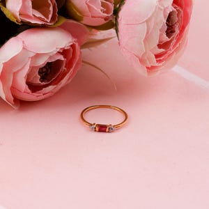 May include: A gold ring featuring a rectangular red gemstone and two small white gemstones. The ring is set against a pink backdrop, with pink flowers in the background. A delicate piece of jewellery.