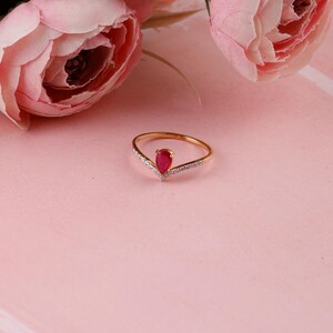 Pu&ograve; includere: Un anello d'oro con una gemma rossa a forma di goccia al centro. L'anello ha un design a forma di V con piccole pietre trasparenti su entrambi i lati della gemma rossa. Fiori rosa sullo sfondo.