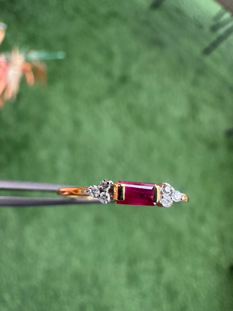 May include: A gold ring with a rectangular, deep red ruby at its centre. The ring is embellished with small, round, clear gemstones clustered on either side of the ruby, creating a delicate and elegant design.