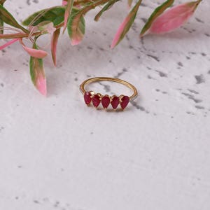 May include: A gold ring featuring five teardrop-shaped, deep red gemstones. The band is gold with small, clear stones set along the top. The ring is displayed on a white surface with pink and green foliage in the background.