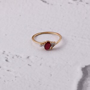 May include: A gold ring with an oval, deep red gemstone at its centre, complemented by smaller, clear stones. The ring has a slender band and is set against a white backdrop.