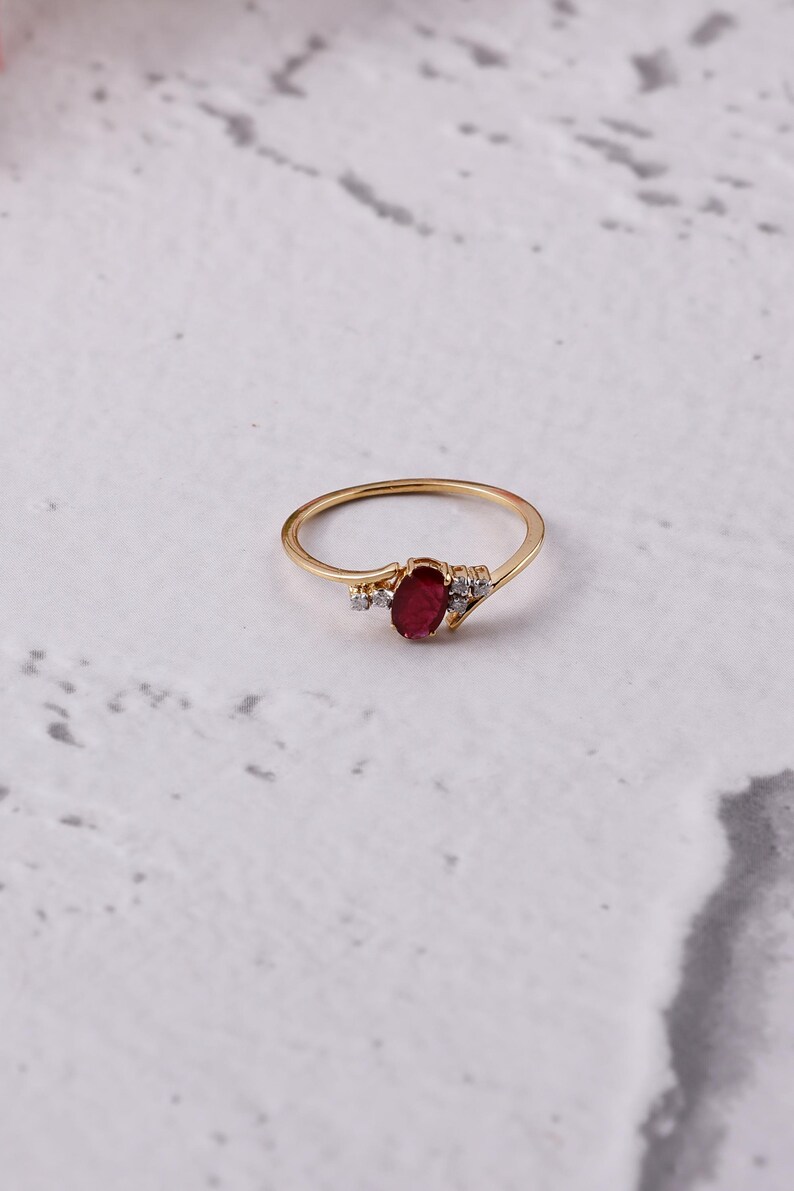 May include: A gold ring with an oval, deep red gemstone at its centre, complemented by smaller, clear stones. The ring has a slender band and is set against a white backdrop.