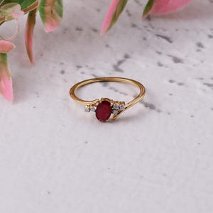 May include: A gold ring with an oval ruby centre stone and small, clear accent stones. The ring has a slender, polished band. The design is delicate and elegant. The ring is displayed on a neutral background, showcasing its refined simplicity.