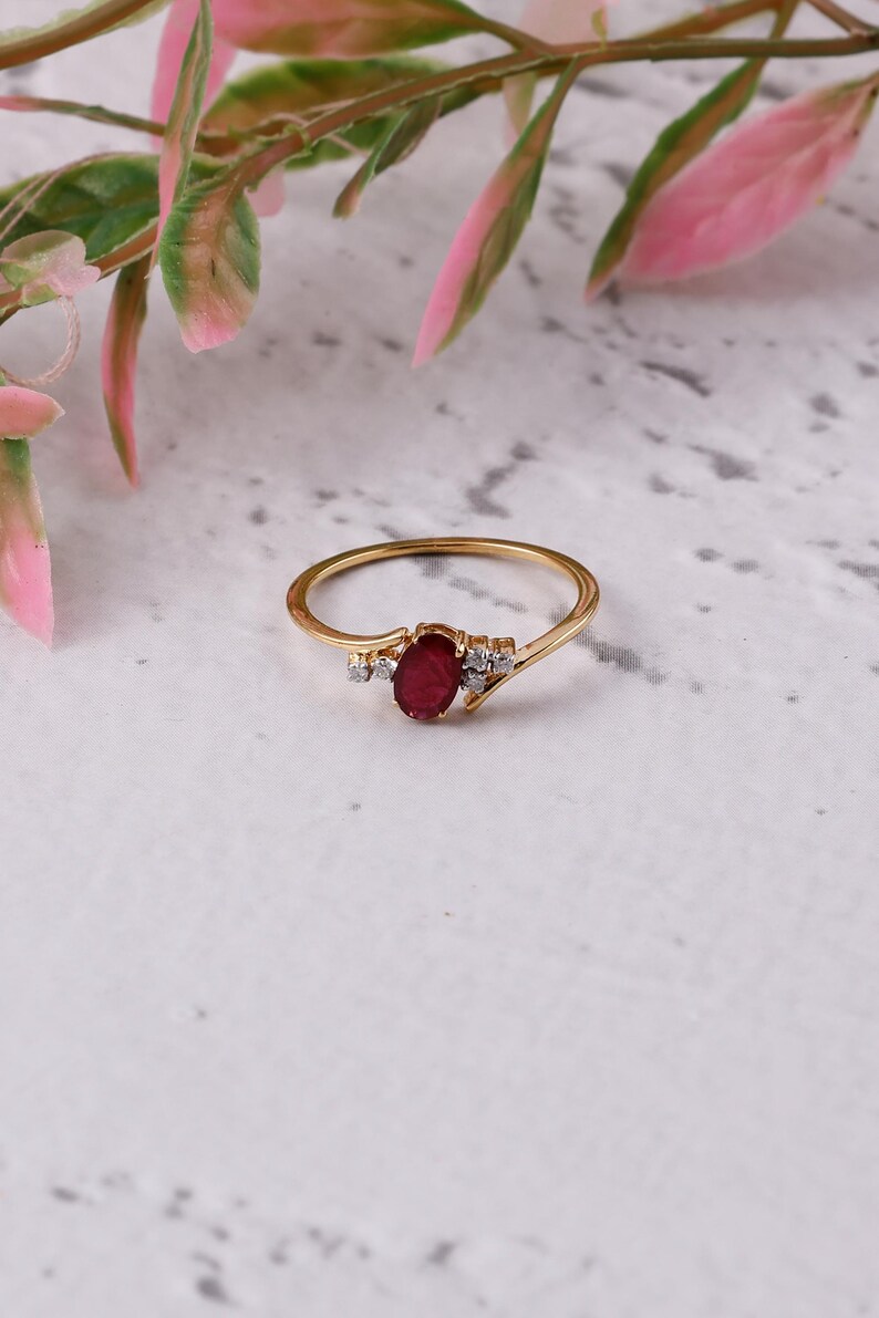 May include: A gold ring with an oval ruby centre stone and small, clear accent stones. The ring has a slender, polished band. The design is delicate and elegant. The ring is displayed on a neutral background, showcasing its refined simplicity.