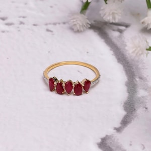 Pode incluir: Um anel de ouro com seis pedras preciosas vermelhas em forma de gota. A fina banda de ouro tem pequenas pedras transparentes na borda superior. O anel &eacute; apresentado em uma superf&iacute;cie de m&aacute;rmore branco e cinza.