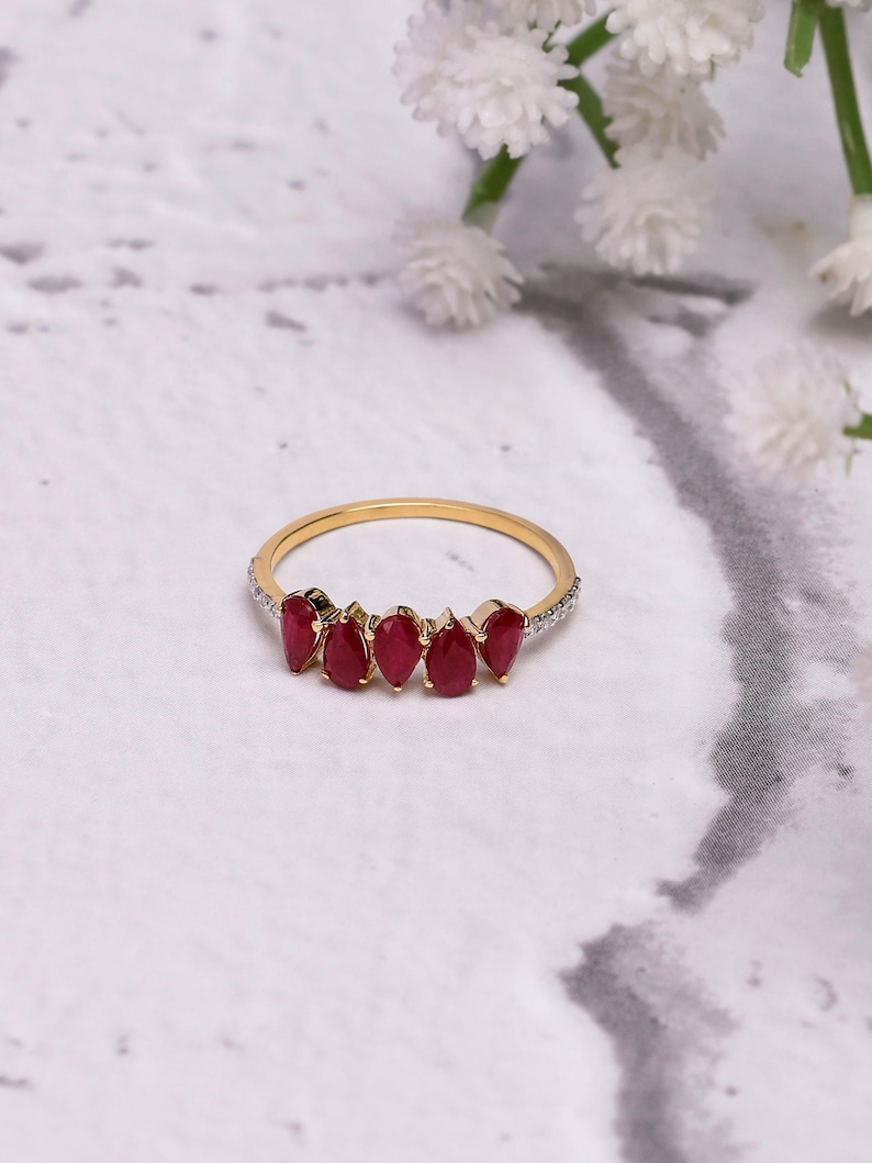 Pode incluir: Um anel de ouro com seis pedras preciosas vermelhas em forma de gota. A fina banda de ouro tem pequenas pedras transparentes na borda superior. O anel &eacute; apresentado em uma superf&iacute;cie de m&aacute;rmore branco e cinza.