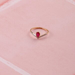 Pu&ograve; includere: Un anello dorato con una gemma rossa a forma di goccia al centro. L'anello ha un design a forma di V con piccole pietre trasparenti lungo la fascia. L'anello &egrave; posto su uno sfondo rosa.