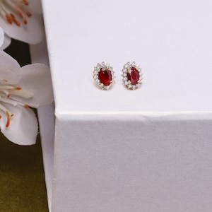May include: A pair of gold-coloured stud earrings, each with a vibrant red oval gemstone surrounded by a halo of small, clear gemstones. The earrings are displayed on a white surface, with a white flower in the background.