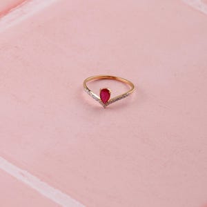 Pu&ograve; includere: Un anello dorato con una gemma rossa a forma di goccia al centro. L'anello ha un design a forma di V con piccole pietre trasparenti lungo la fascia. L'anello &egrave; posto su uno sfondo rosa.