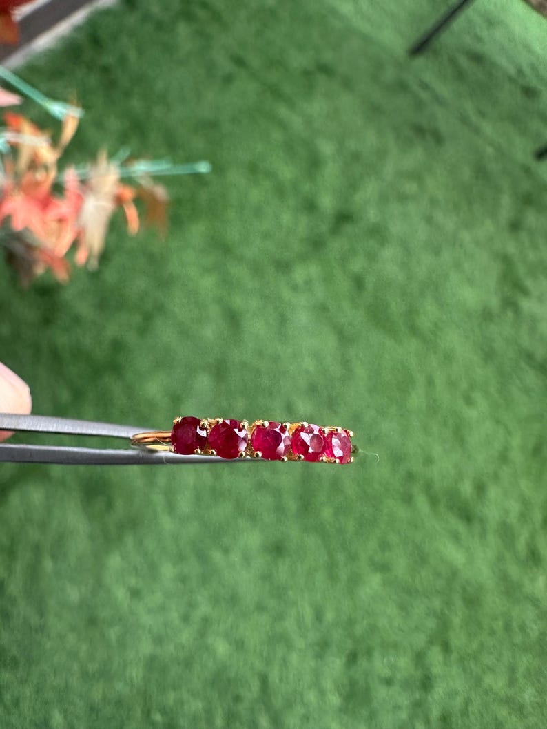 Op de afbeelding: Een gouden ring met vijf ovaal geslepen, dieprode edelstenen. De ring wordt door een pincet tegen een groene achtergrond gehouden. De edelstenen zijn gezet in een gouden band, met een klassiek ontwerp.