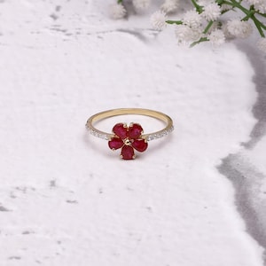 May include: A gold ring with a flower-shaped cluster of six red gemstones. The band is embellished with small, clear stones. The ring is set against a white backdrop with sprigs of white flowers.
