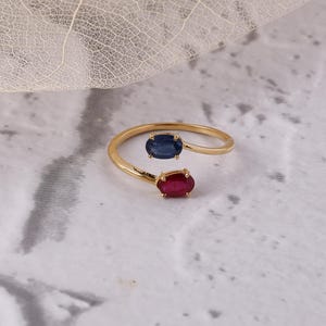 May include: A gold ring with a bypass design, set with two oval gemstones. One is a deep blue, the other a vibrant red. The ring sits on a marbled surface. The gemstones are approximately 0.5 cm in length.