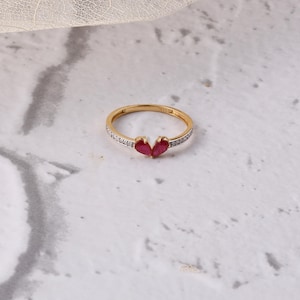 May include: A gold ring with a heart-shaped design. The ring features two red gemstones at the centre and small white gemstones along the band. The ring is set against a white and grey marble background.