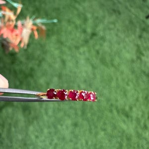 Op de afbeelding: Een gouden ring met vijf levendige rode edelstenen. De edelstenen zijn in een rij gerangschikt, waardoor een doorlopende band van kleur ontstaat. De ring wordt door een pincet tegen een groene achtergrond gehouden.