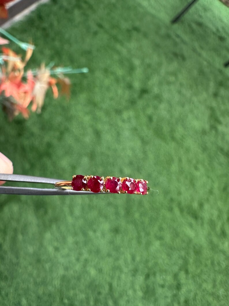 Op de afbeelding: Een gouden ring met vijf levendige rode edelstenen. De edelstenen zijn in een rij gerangschikt, waardoor een doorlopende band van kleur ontstaat. De ring wordt door een pincet tegen een groene achtergrond gehouden.