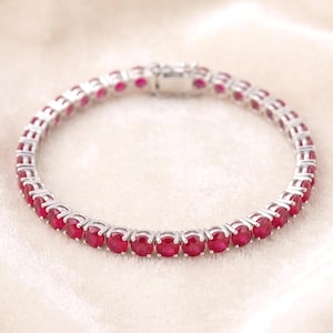 May include: A silver-toned tennis bracelet featuring a continuous row of round, vibrant red gemstones set in individual metal prongs. The bracelet includes a secure, rectangular box clasp mechanism at the top, creating a circular shape.