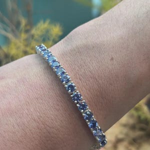 Può includere: Un bracciale tennis caratterizzato da una fila continua di pietre preziose rotonde color blu pervinca, incastonate in una fascia di metallo color argento lucido. Le pietre sono uniformi per dimensioni e forma, creando un design lineare luminoso.